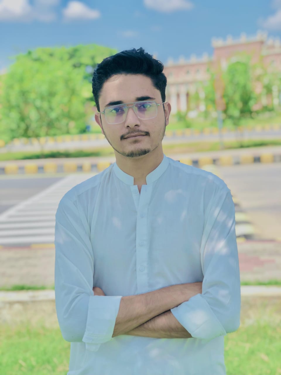 A person in a white button-up shirt stands with arms crossed in a sunlit park, with a blurred historic building and lush green trees in the background.