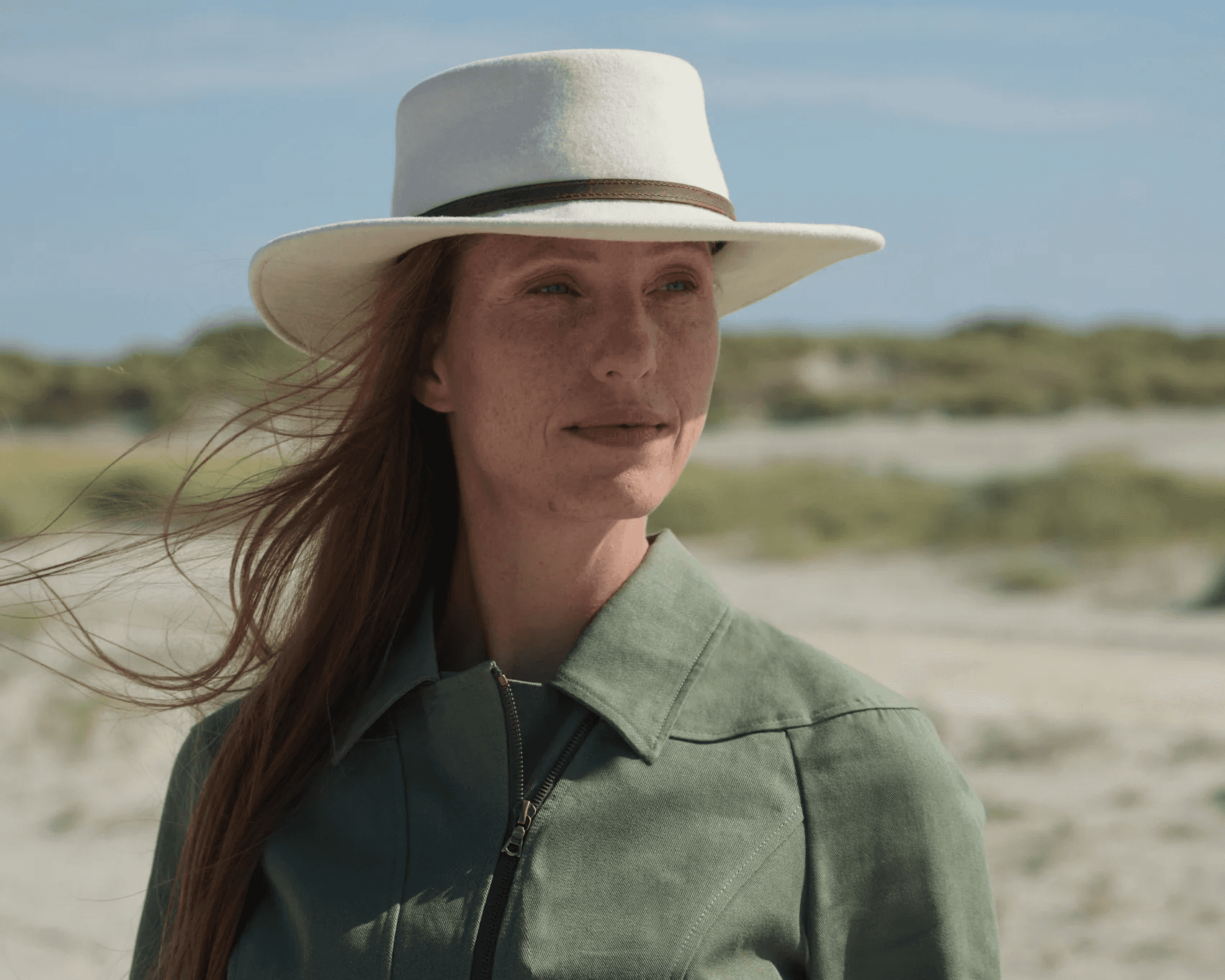 A woman with long hair wears a wide-brimmed white hat and a green jacket, standing in a sunny, open landscape with blurred greenery and sand in the background.