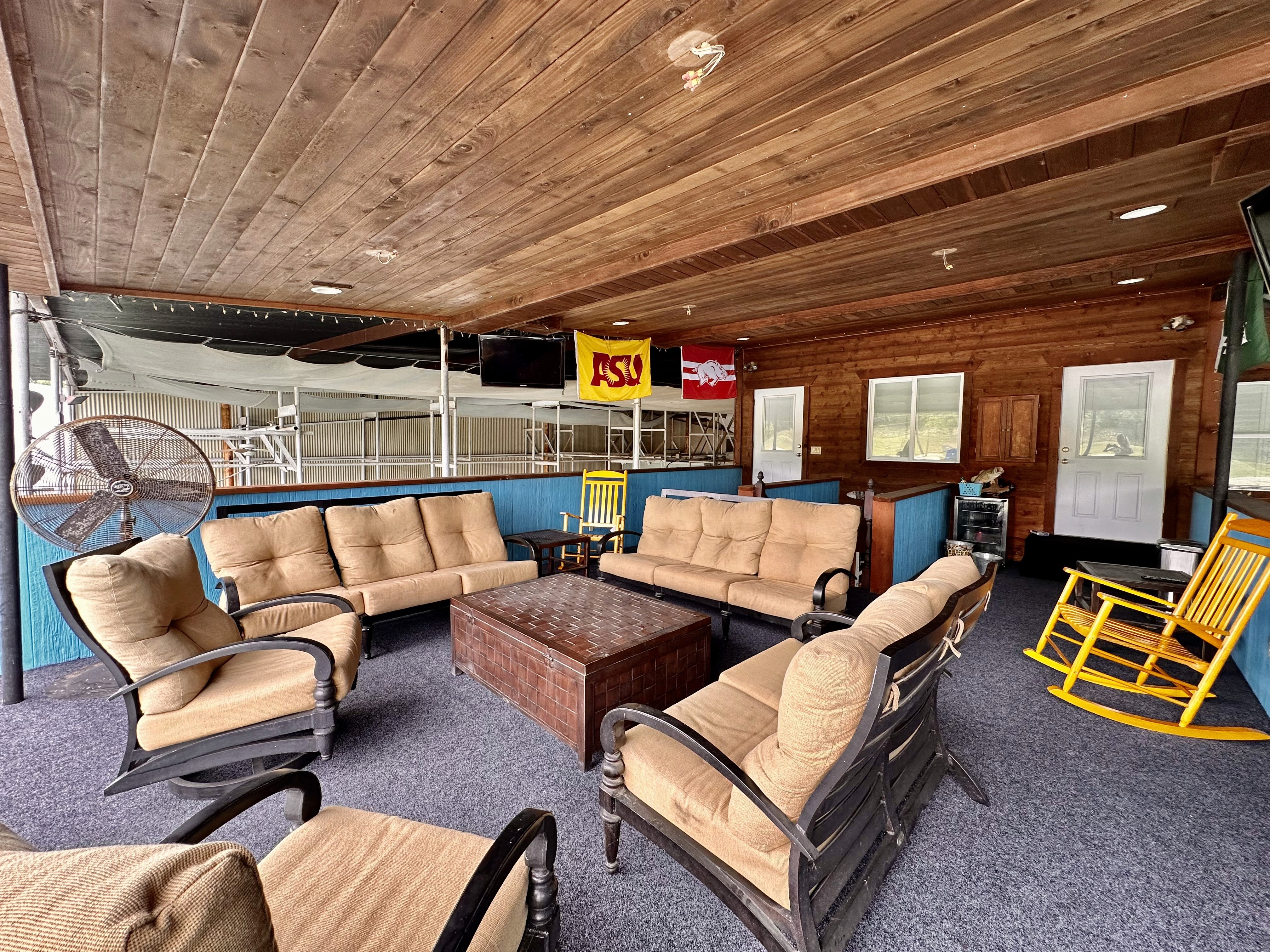 A cozy, covered patio features beige cushioned seating arranged around a central wooden table, with vibrant yellow rocking chairs and colorful college flags adding a touch of character under a wooden ceiling.