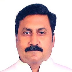 A smiling person with curly hair sits at a desk, looking at a laptop against a dark background.