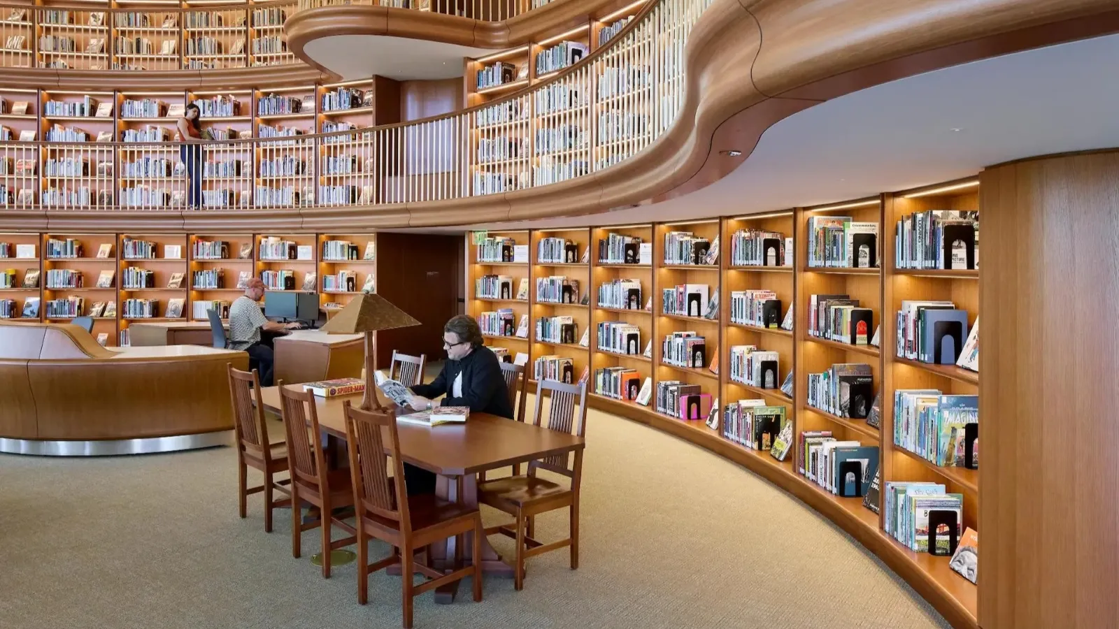 A modern library interior featuring curved wooden bookshelves filled with books, a long table with chairs in the foreground, and individuals engaged in reading and working on laptops.