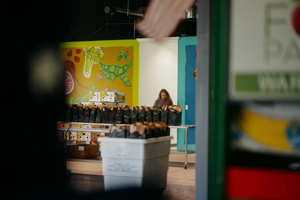 A photograph taken indoors, slightly framed by a dark foreground on the left, shows a woman standing behind a long table. She is looking down and organizing a long row of black bags, which appear to be filled with groceries or supplies. The walls are decorated with large, colorful murals of produce, including a yellow one with illustrations of vegetables and a blue-green one with partially visible text on the right. A large white plastic storage crate is visible in the foreground on the bottom right. The scene suggests an operation at a food bank or distribution center.