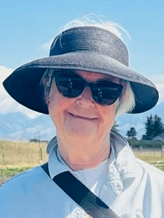 A woman with shoulder-length brown hair, wearing a white shirt, smiles against a blurred outdoor background.