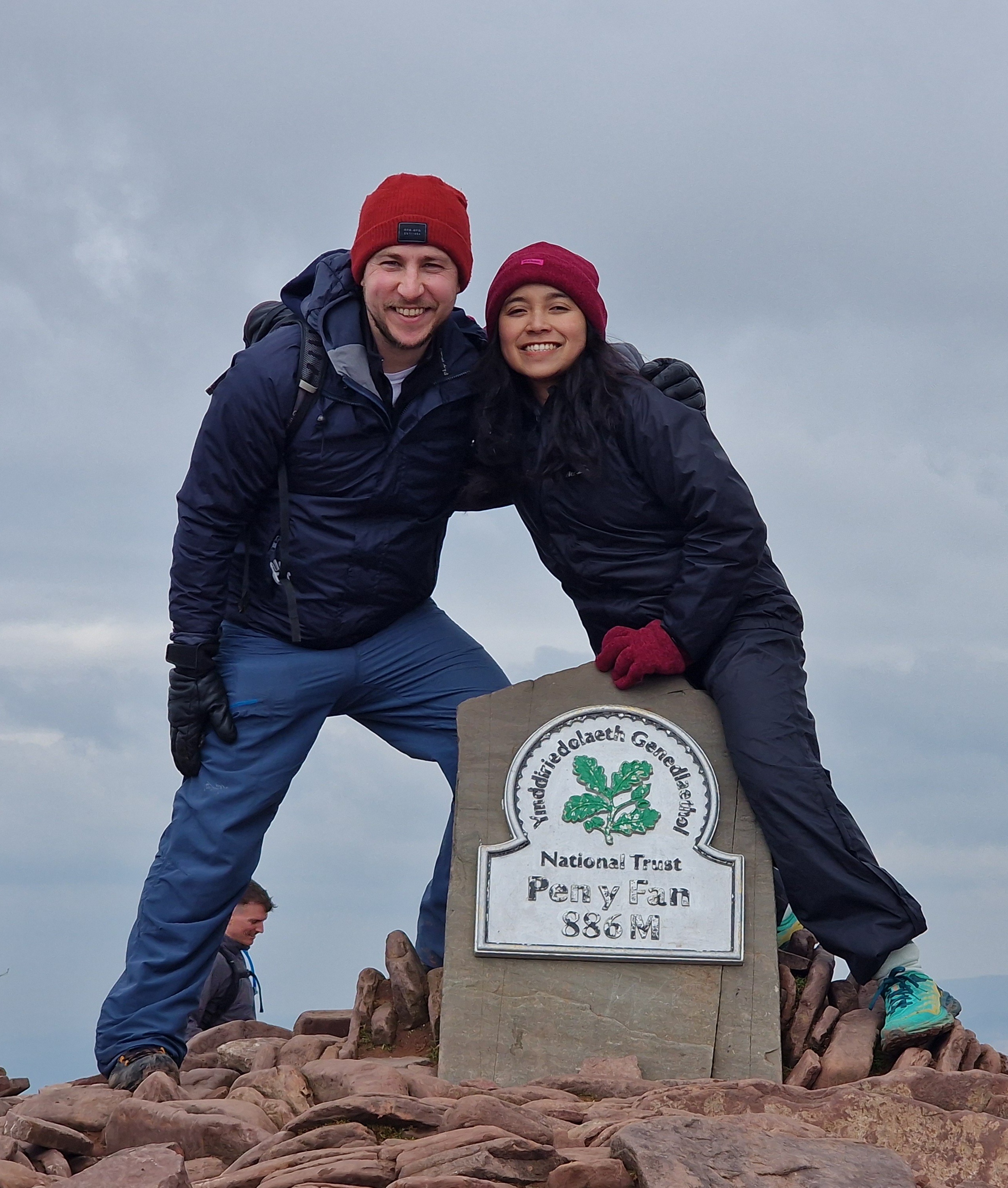 A woman with long blonde hair, in a brown top and jeans, walks through a sunlit forest, holding someone's hand. The scene is warm and inviting.