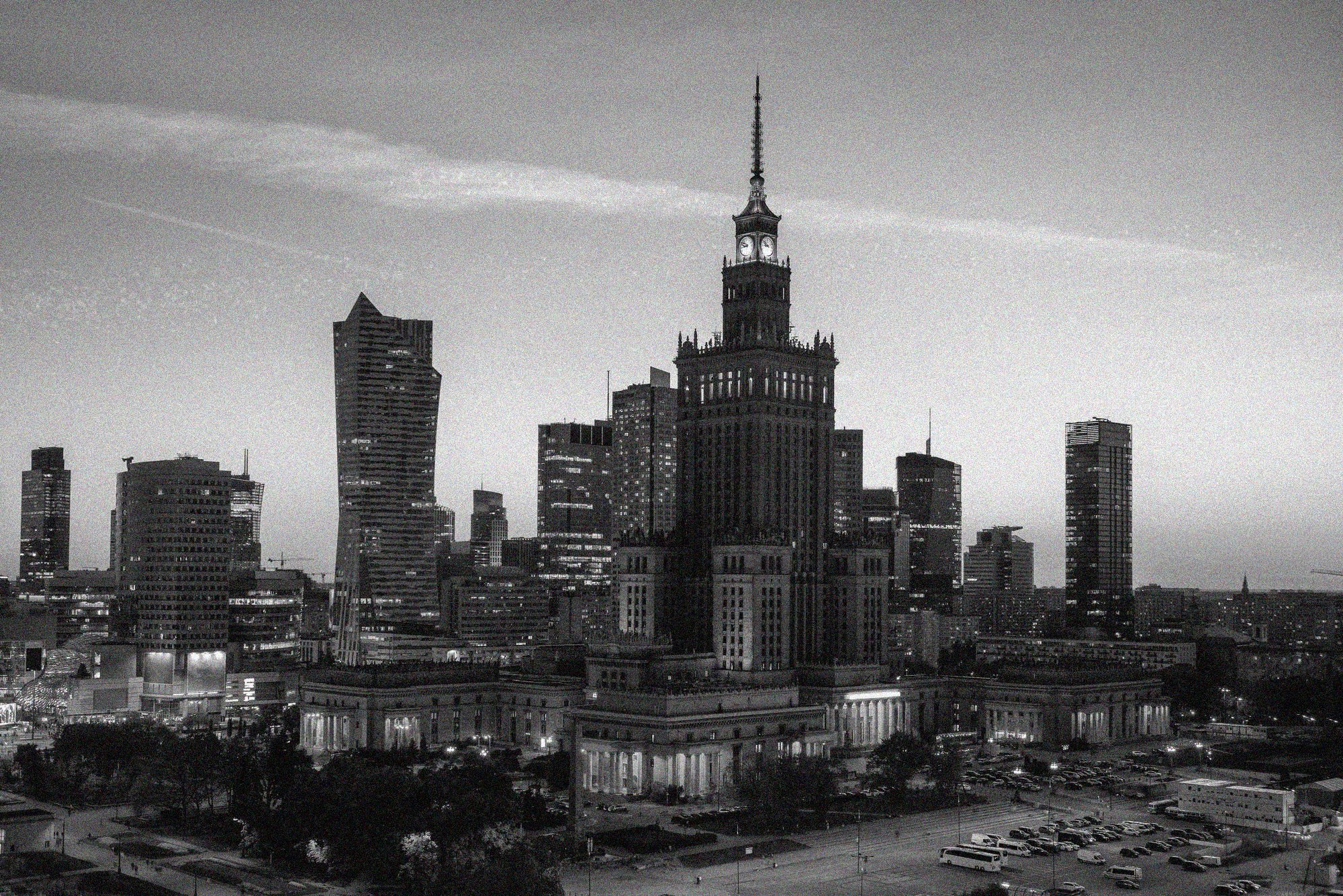 Black-and-white Warsaw skyline with the Palace of Culture and Science rising above modern high-rise buildings at dusk. 