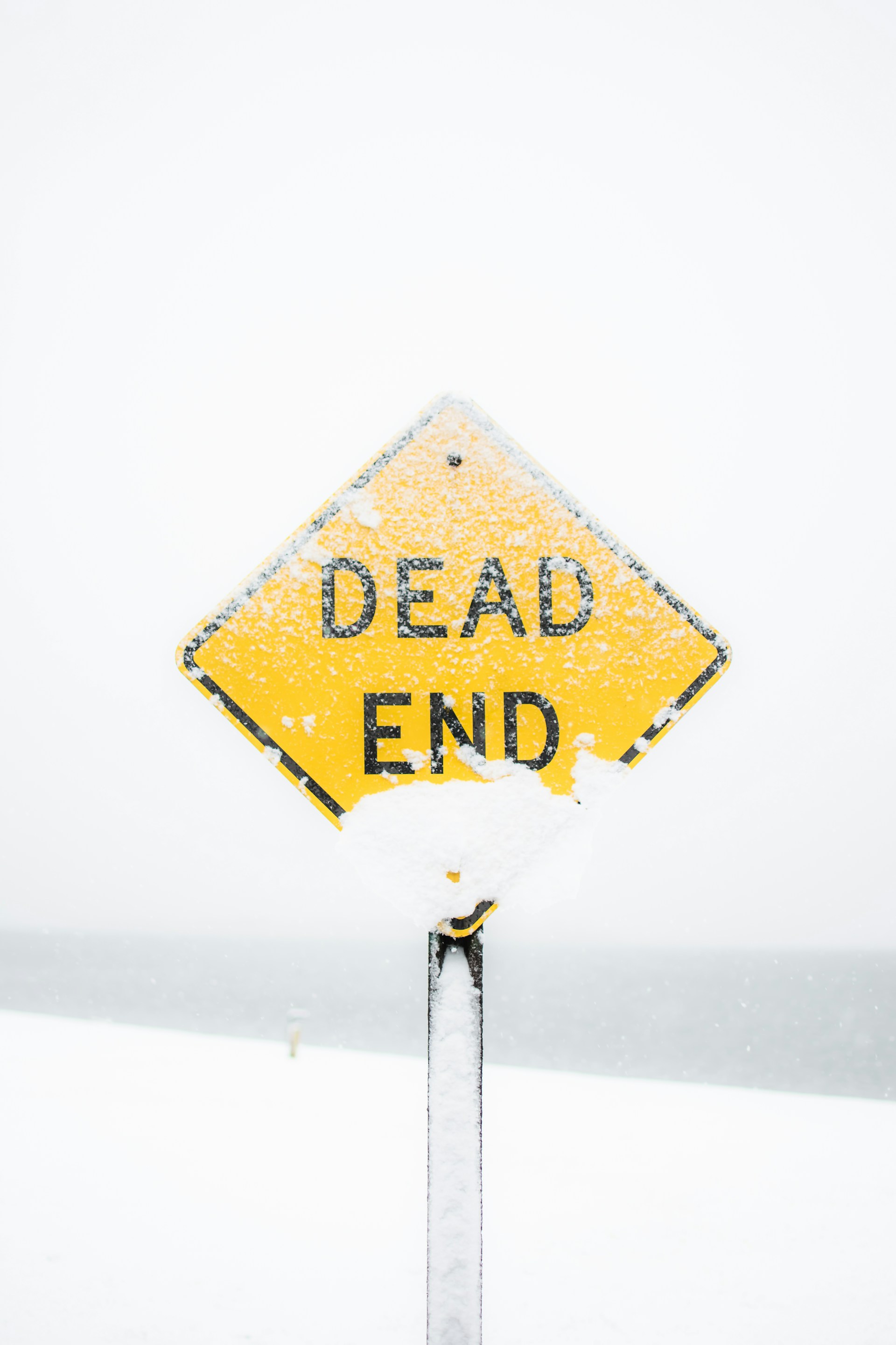 Yellow "Dead End" road sign partially covered in snow, standing against a snowy, white winter landscape with a hazy, overcast sky in the background