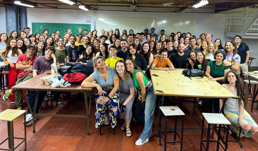 A person wearing headphones sits at a desk with a large curved monitor displaying graphic design software, surrounded by a laptop, smartphone, and potted plant, creating a modern workspace setup.