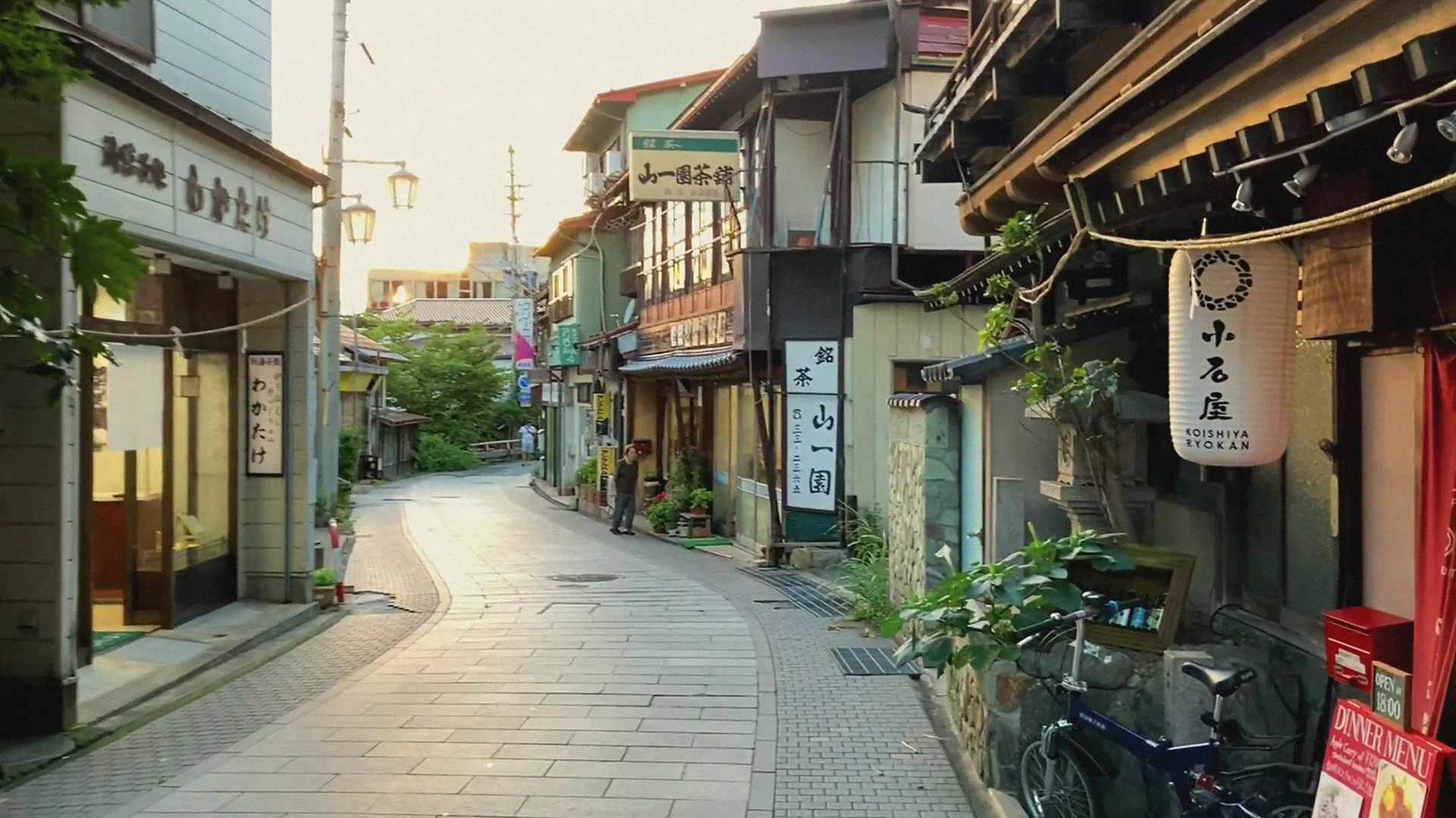Shibu Onsen's curving stone-paved lane at golden hour — wooden ryokan, paper lanterns and a Koishiya inn sign in warm light.