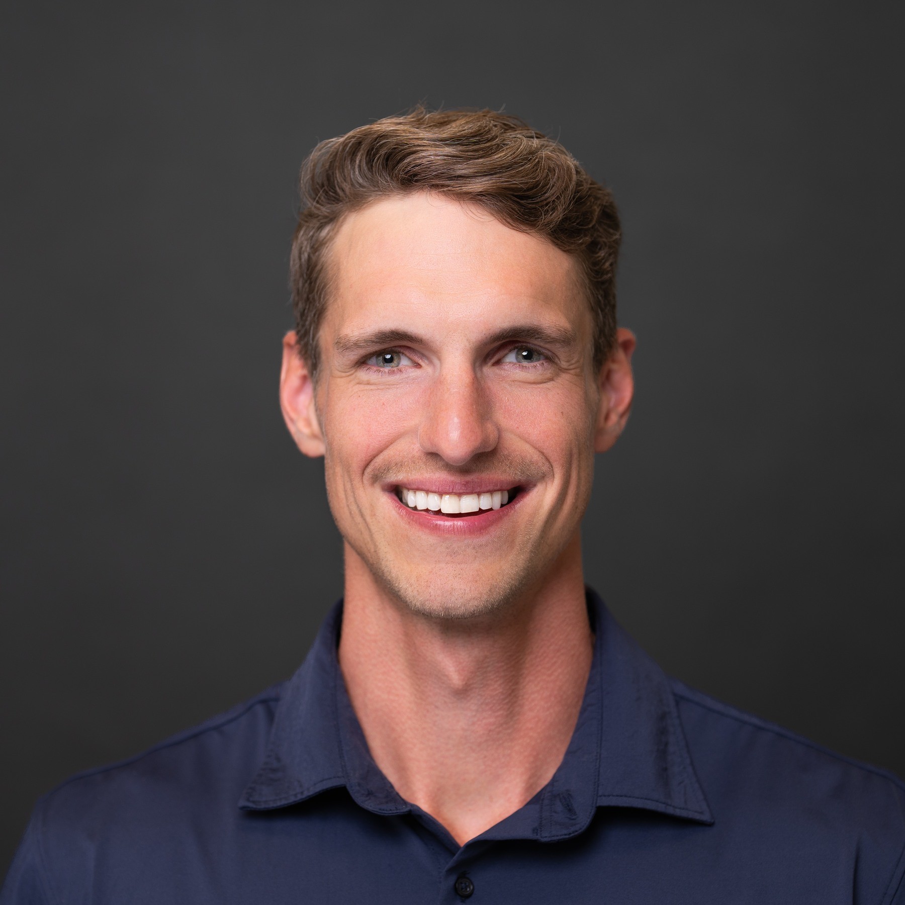 Man in a navy shirt smiling for a headshot against a dark gray background