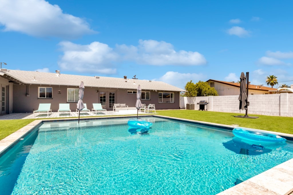 A spacious backyard features a sparkling swimming pool with two inflatable loungers, surrounded by a well-manicured lawn and modern outdoor seating, showcasing an affordable luxury vacation rental house under a clear blue sky.
