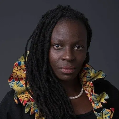 A person with long, braided hair wearing a colorful patterned garment and a pearl necklace, set against a dark gray background.