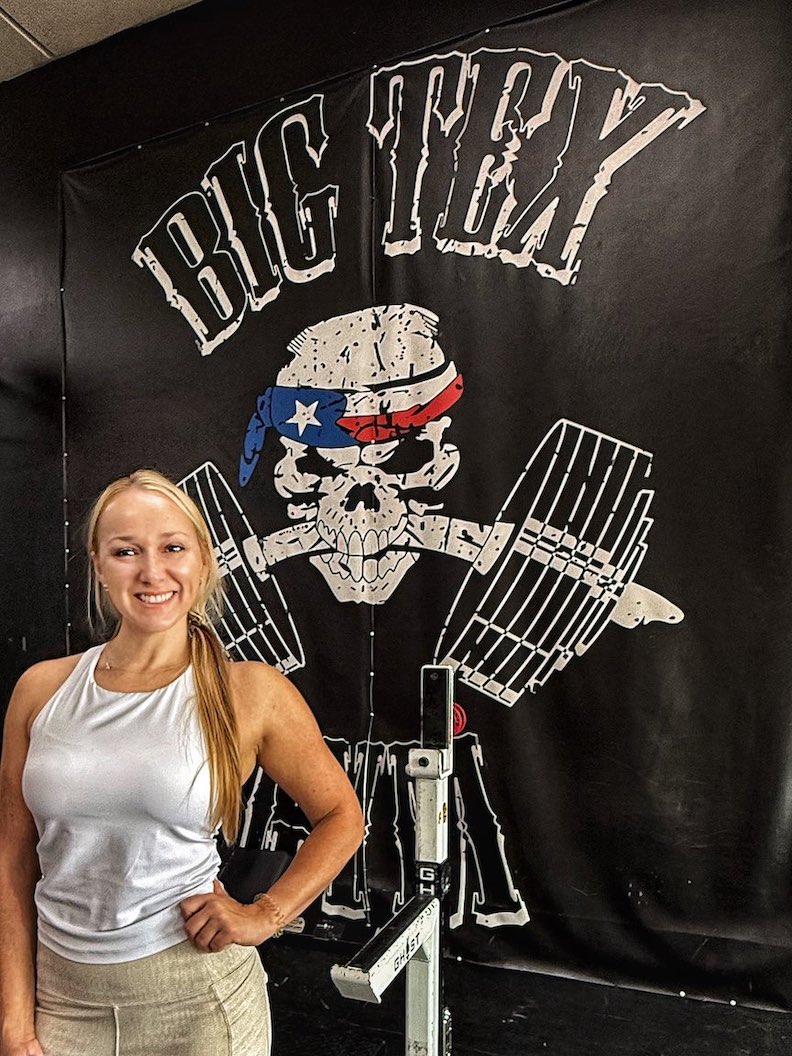 Kathryn Alexander, a trainer at big tex gym, posing in front of the big tex gym poster