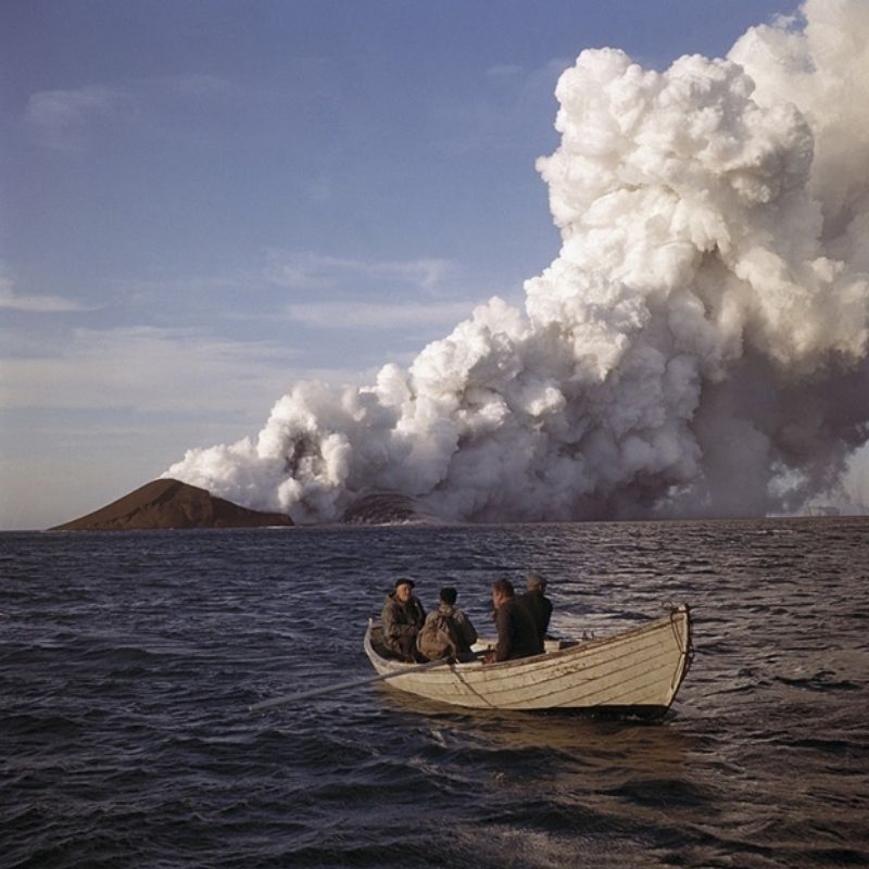 Three men in a row boat form a film crew and approach the erupting volcano of Surtsey, part of the Vestmannaeyjar volcanic system, in hopes to document the new island and the lava.