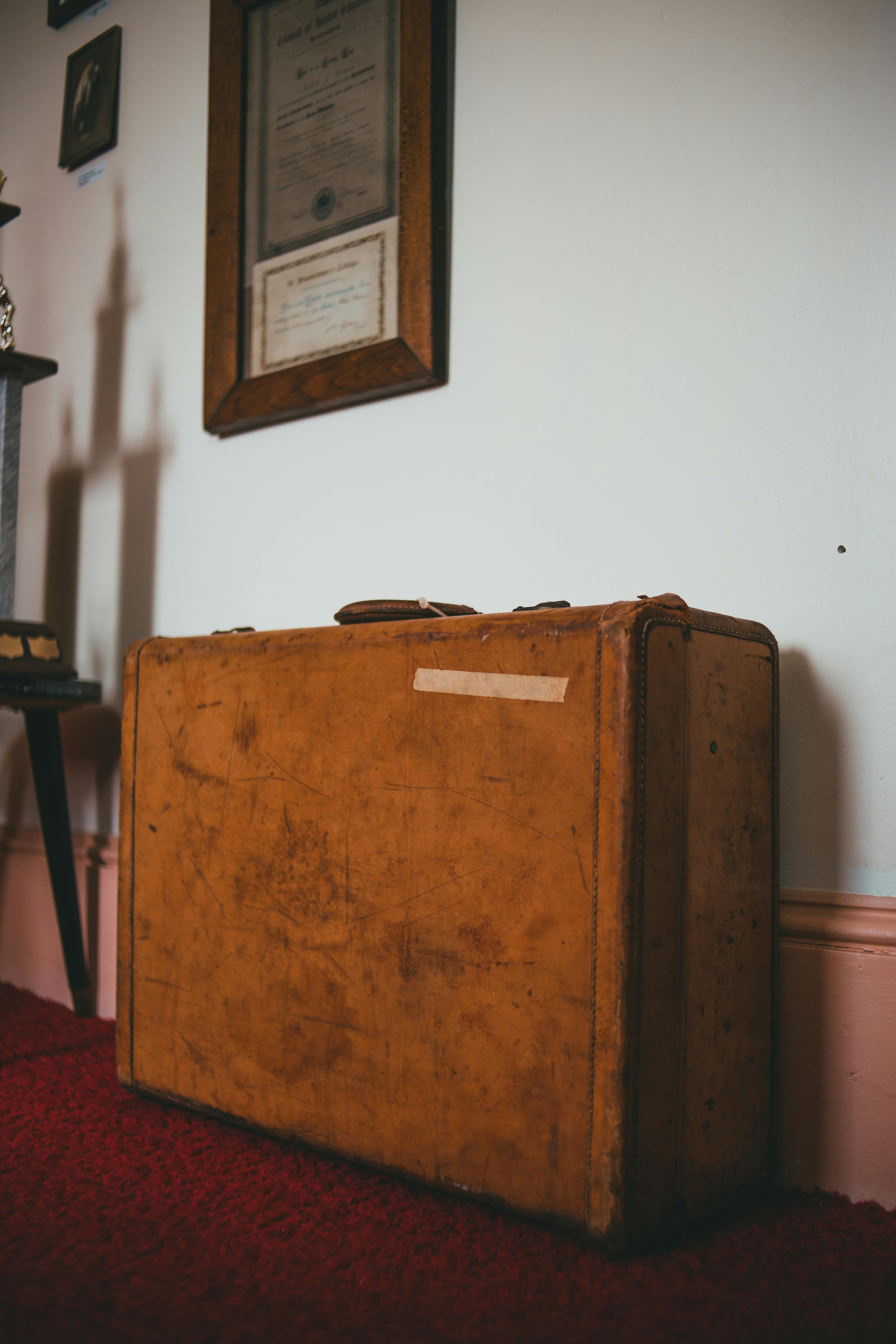A vintage brown leather suitcase rests against a white wall adorned with framed certificates, placed on a red carpeted floor, evoking a nostalgic and timeless atmosphere.