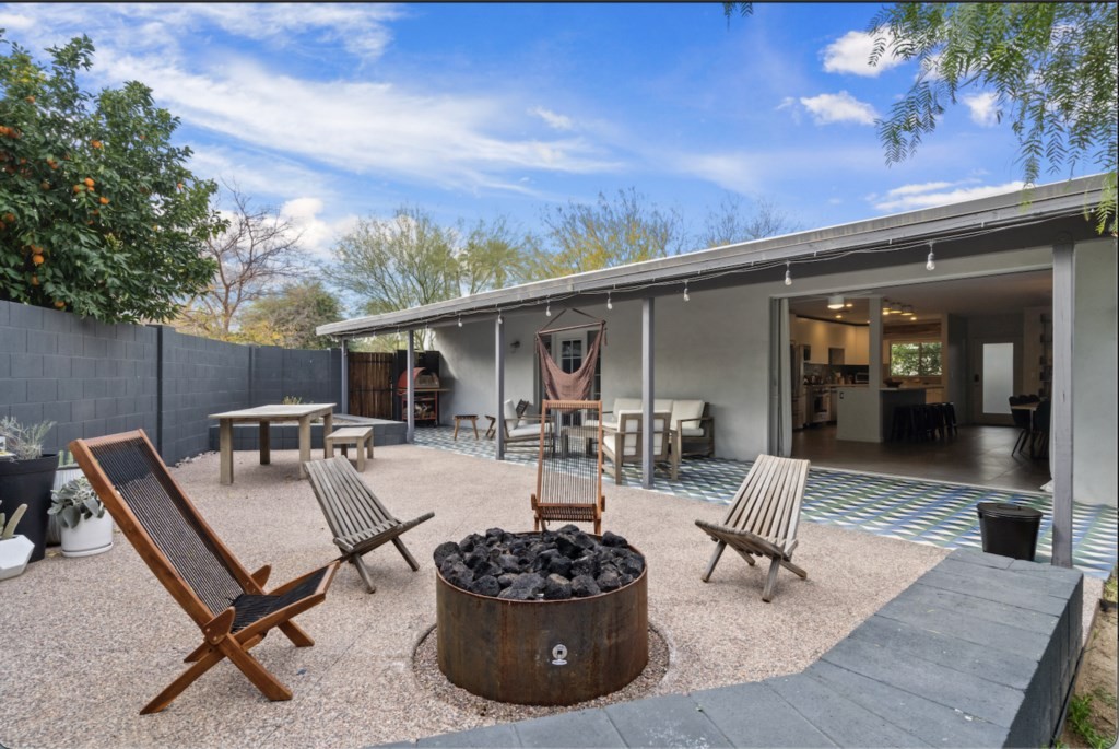 A modern backyard features a cozy fire pit surrounded by stylish wooden chairs, set against a backdrop of a sleek, white affordable luxury vacation rental house with a spacious patio and vibrant tiled flooring.