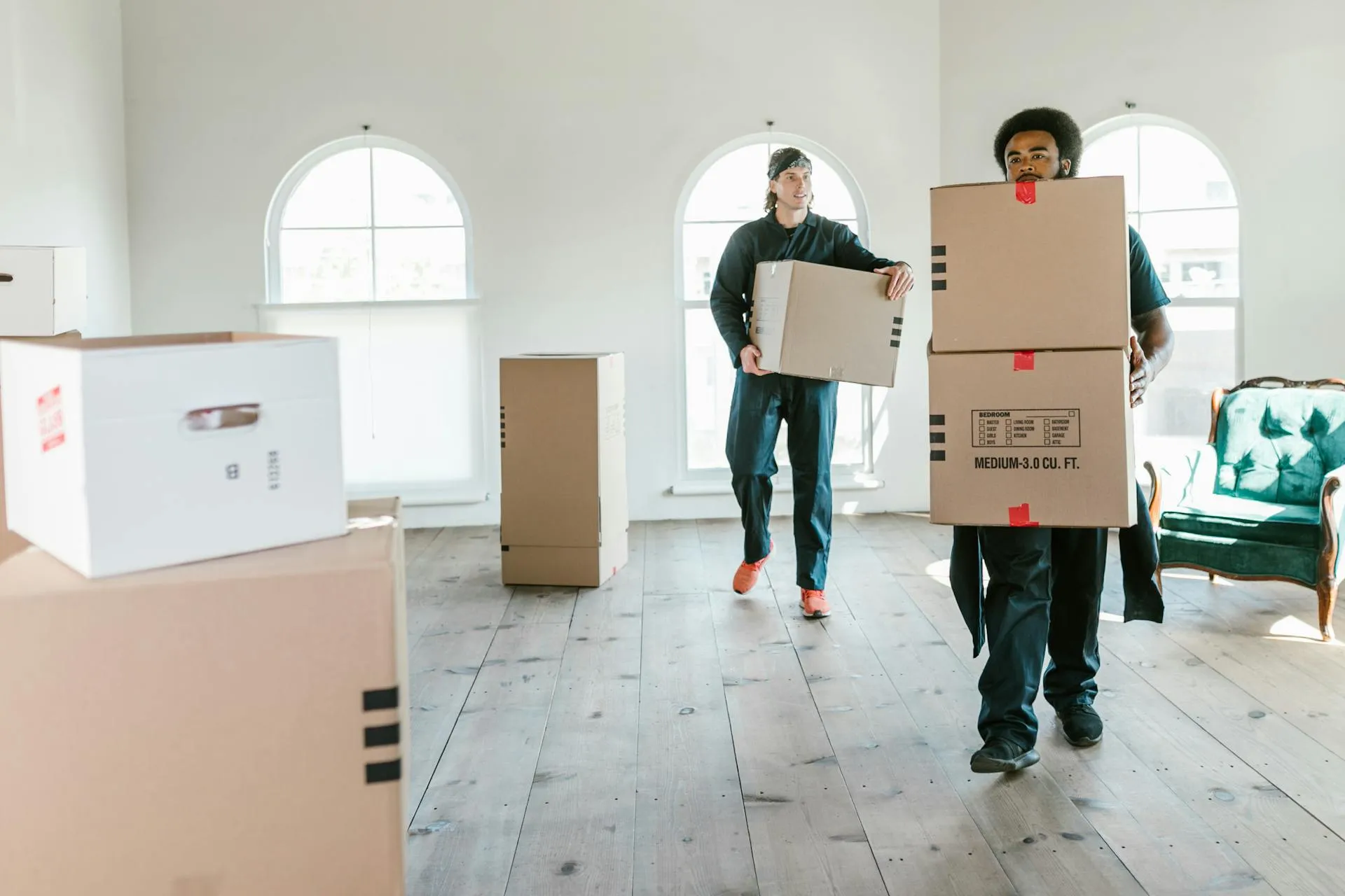 Professional movers carrying heavy cardboard boxes inside a spacious house with large arched windows and wooden floors.