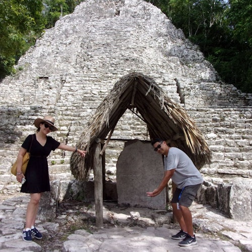 Two people posing near a historical stone monument with a thatched roof covering. A large stone pyramid is in the background.