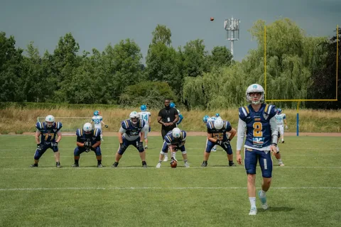 A group of American football players in blue and white uniforms set up on a grassy field, with a quarterback preparing to pass while teammates line up in formation under a cloudy sky.