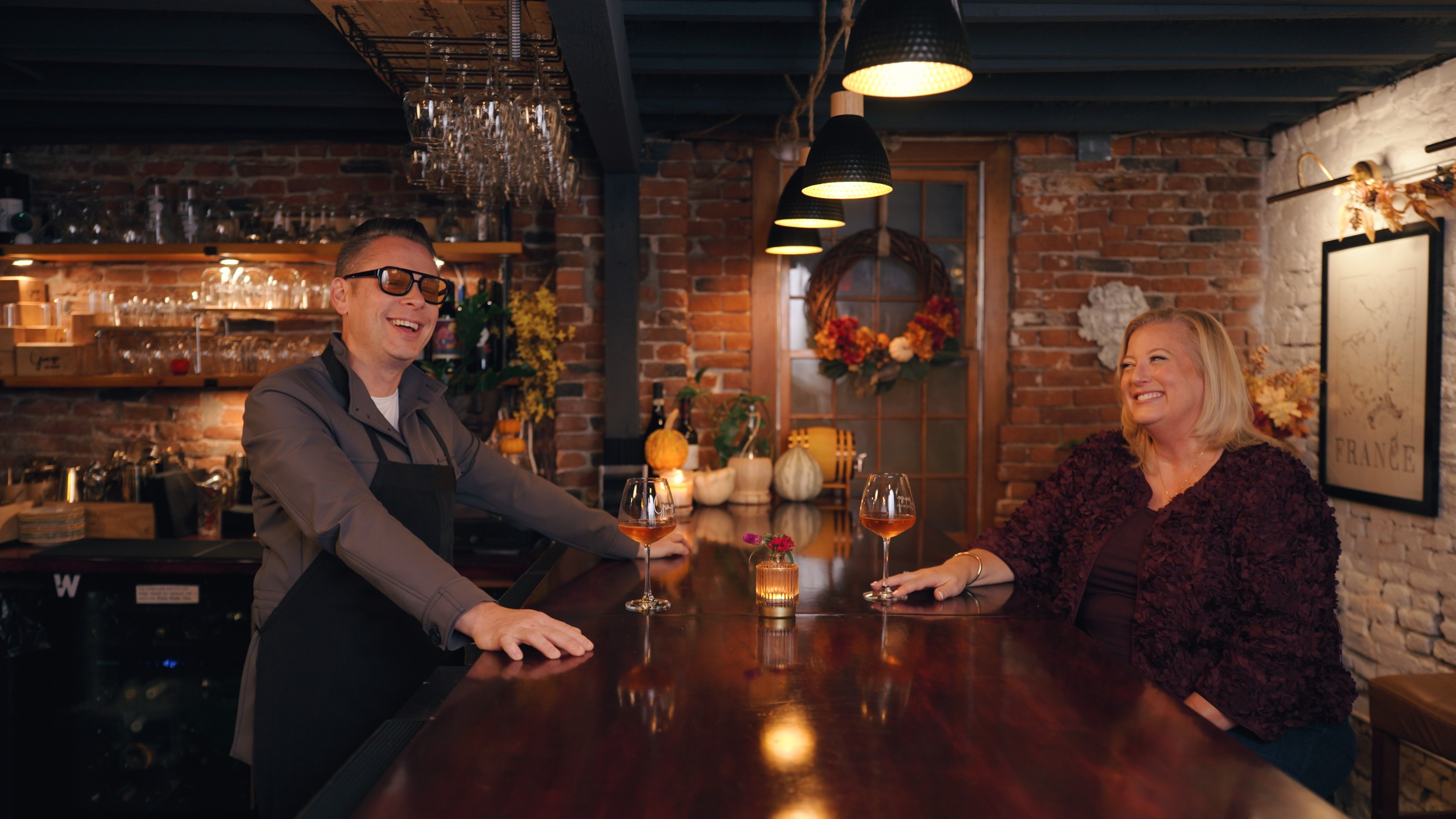 A warm, inviting shot inside a rustic bar or tavern setting featuring two people interacting across a long wooden bar top. On the left, a smiling man with short hair and tinted glasses wears a grey button-down shirt over a black apron, leaning against the bar. On the right, a blonde woman in a textured burgundy blouse sits at the bar, laughing and looking toward the man. Between them on the polished wooden counter sit two glasses of amber-colored wine and a small glowing candle holder. The background shows exposed brick walls, hanging glassware, decorative gourds, an autumnal door wreath, and black pendant lights casting a cozy glow over the scene.
