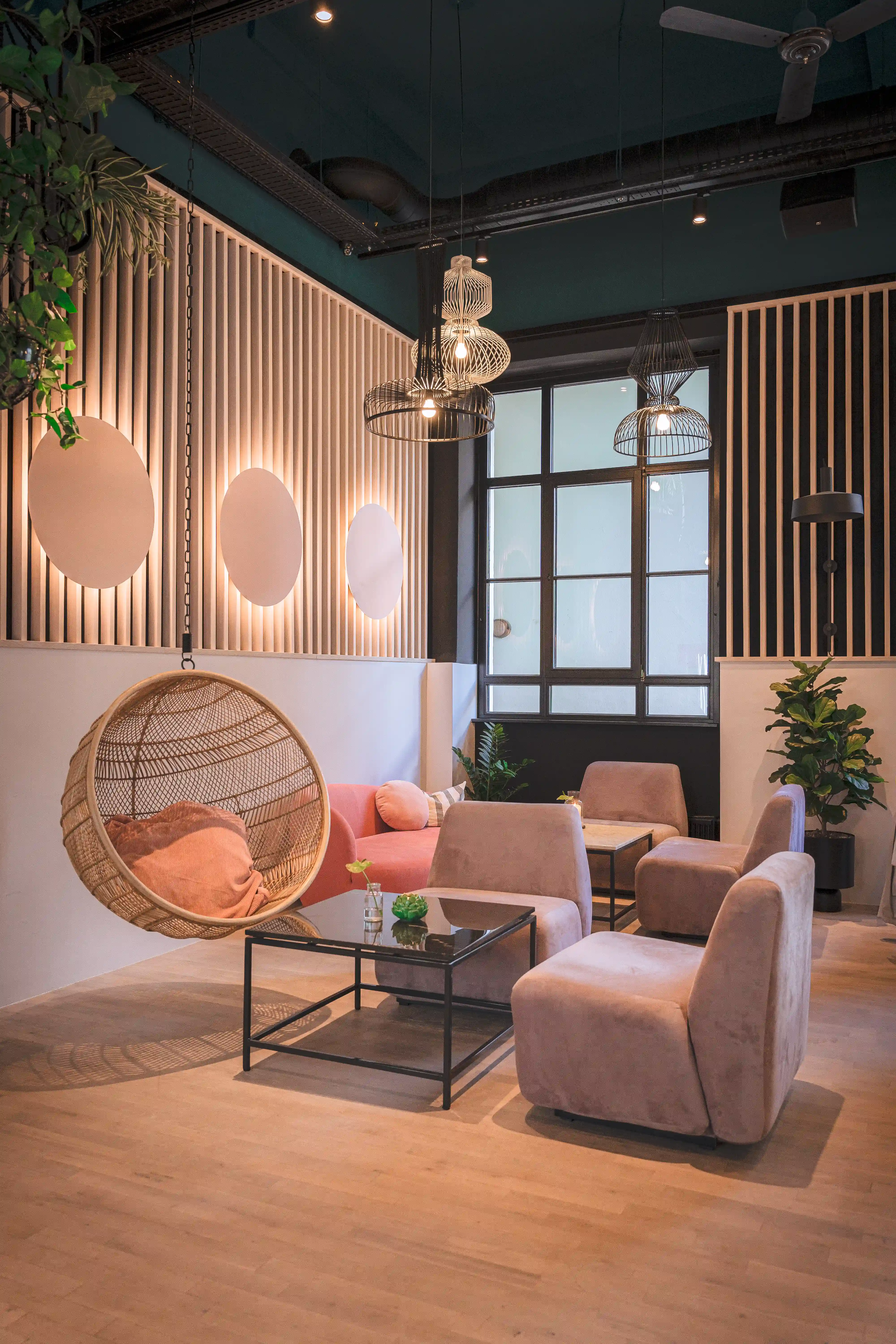 Cosy corner at LOKAL VIER with a hanging rattan chair with pink cushions, blush velvet armchairs, a glass coffee table and glowing circles on slatted wall panels.