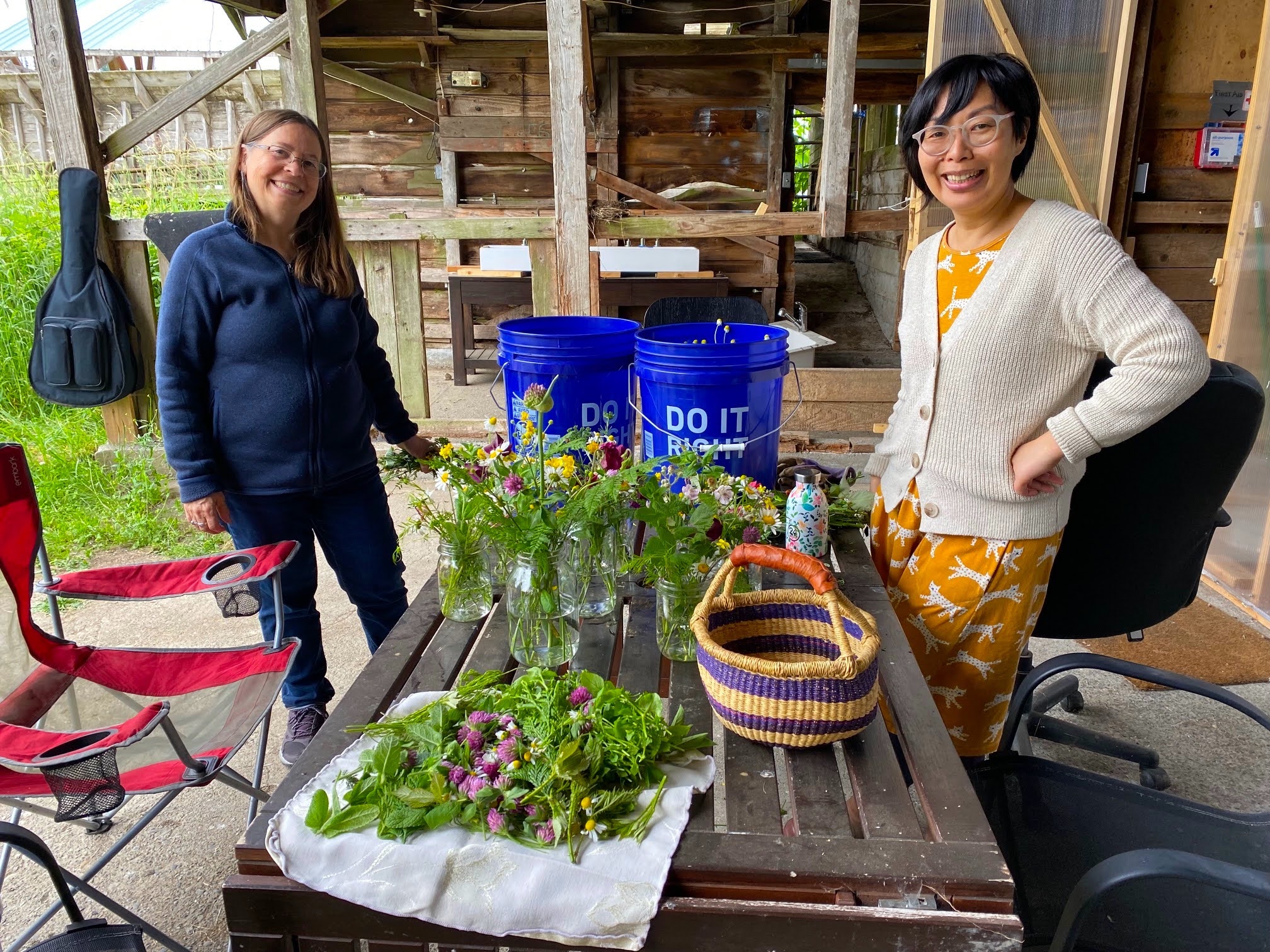 Two people standing beside a table with garden seedlings, baskets, and blue bins in a rustic barn-like space.