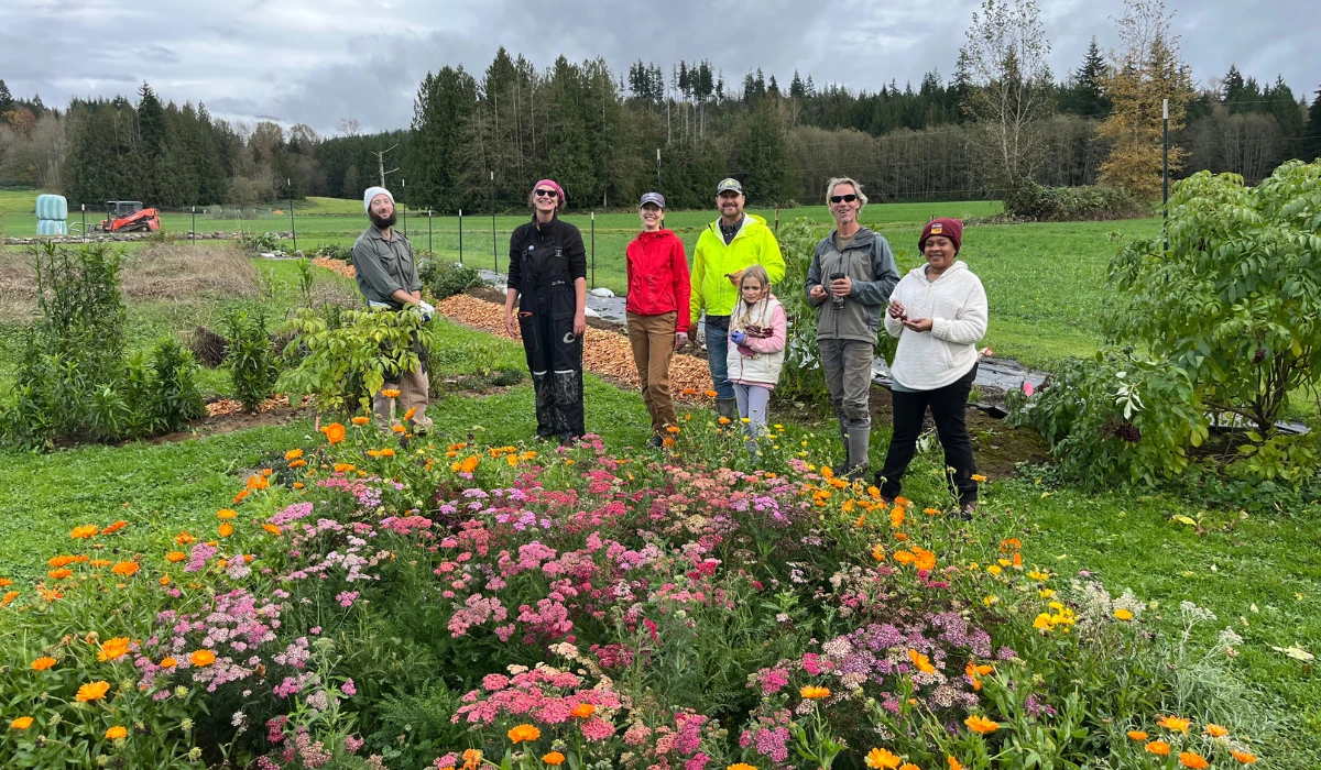Community members stand together in a garden at Rooted Northwest, surrounded by blooming flowers and farmland.