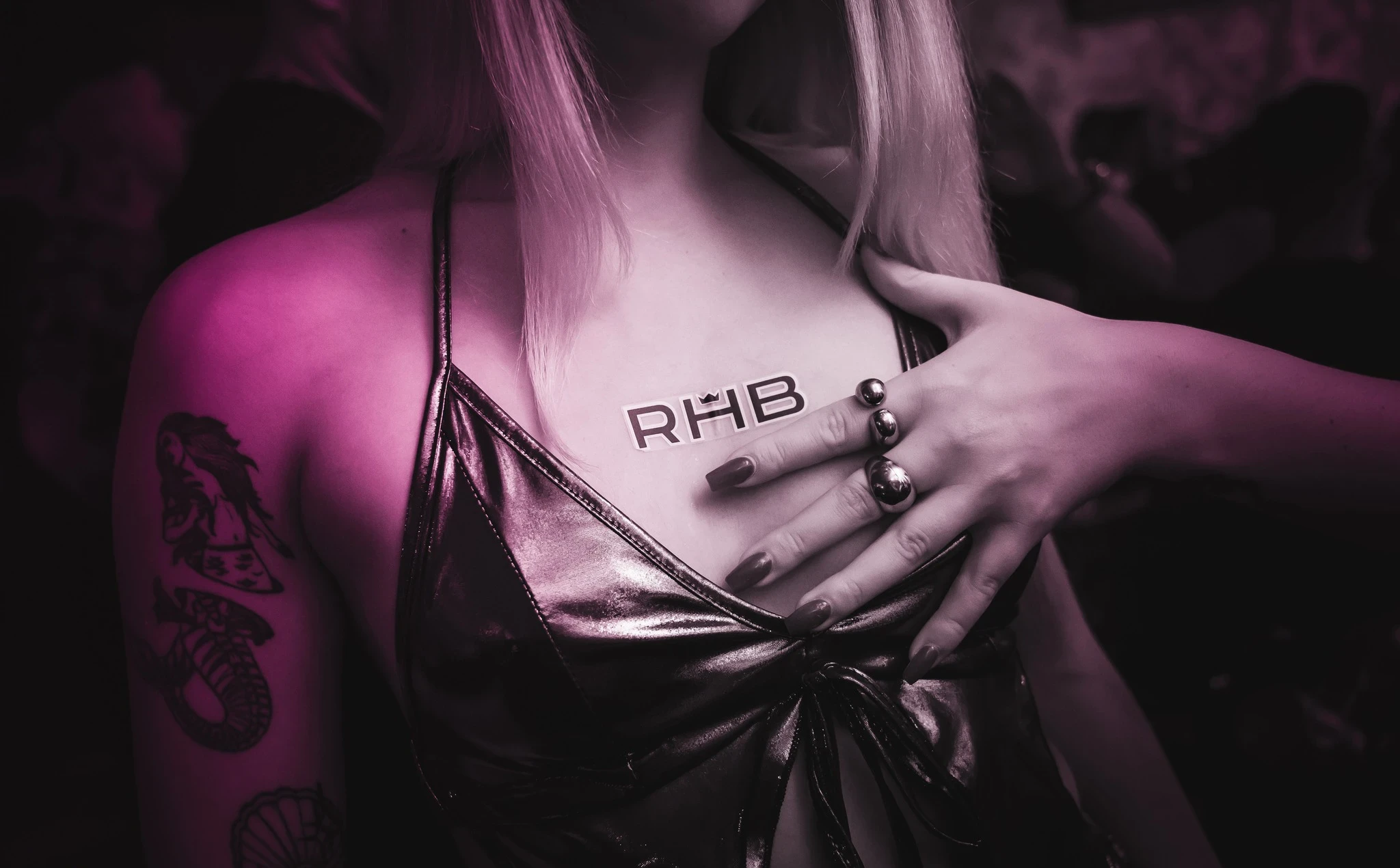 A close-up shot focusing on a person's chest and shoulder at the RHB party. They wear a metallic top, have a large mermaid tattoo, and another person's hand rests on their chest near an "RHB" sticker.