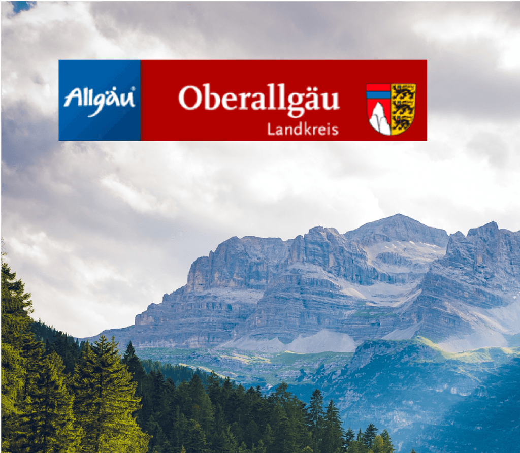Im Vordergrund ein Waldabschnitt mit großen Tannen hinter dessen sich ein großer Berg erstreckt mit einer riesigen Wolkendecke darüber darauf das Logo des Landkreis Oberallgäu