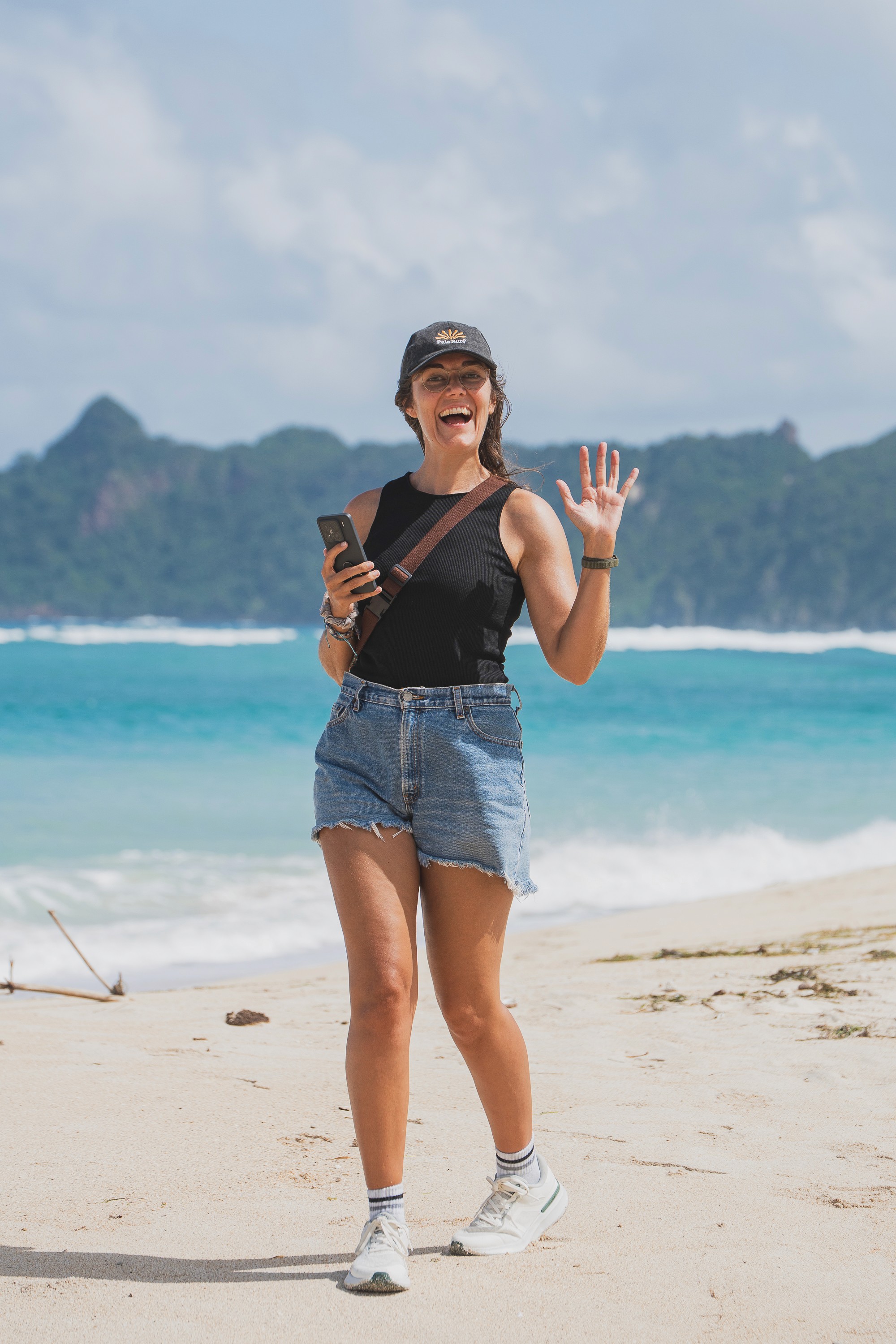 Eva smiling while walking on a sandy beach beside clear blue water.