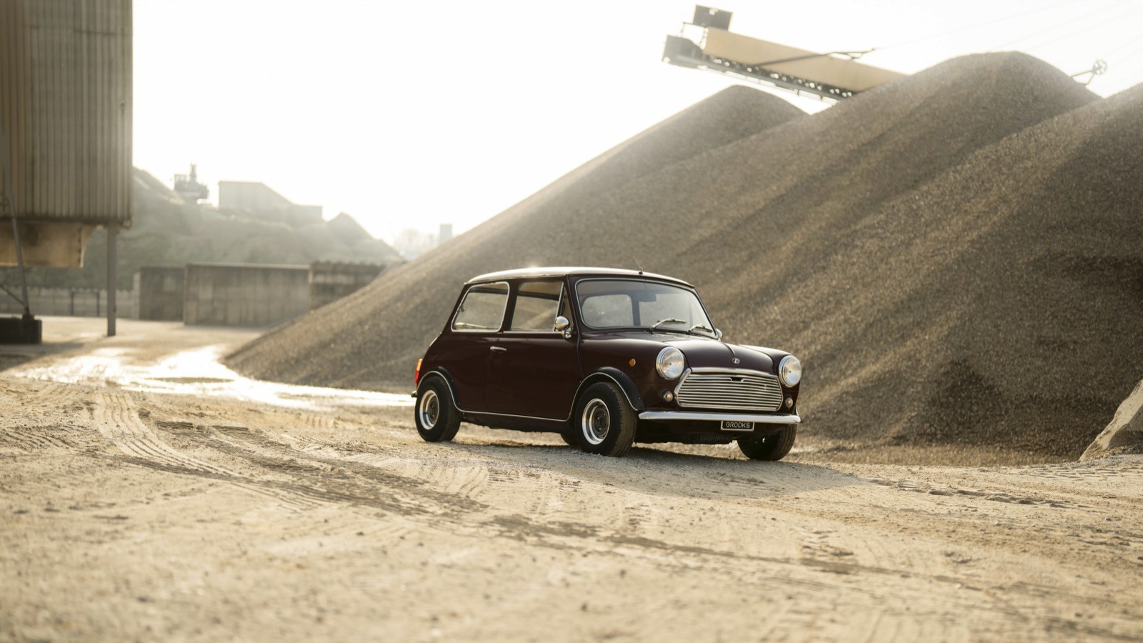 A vintage car parked on a sandy surface with large piles of sand in the background under a clear sky.