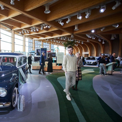 Personer som går i ett modernt showroom med vintage- och moderna bilar utställda under träbjälkar och ljus belysning.