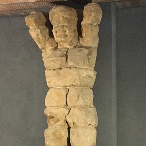 一座由堆疊不規則石塊製成的雕塑，形似舉起雙臂的人形，靠在一面平坦的牆上。