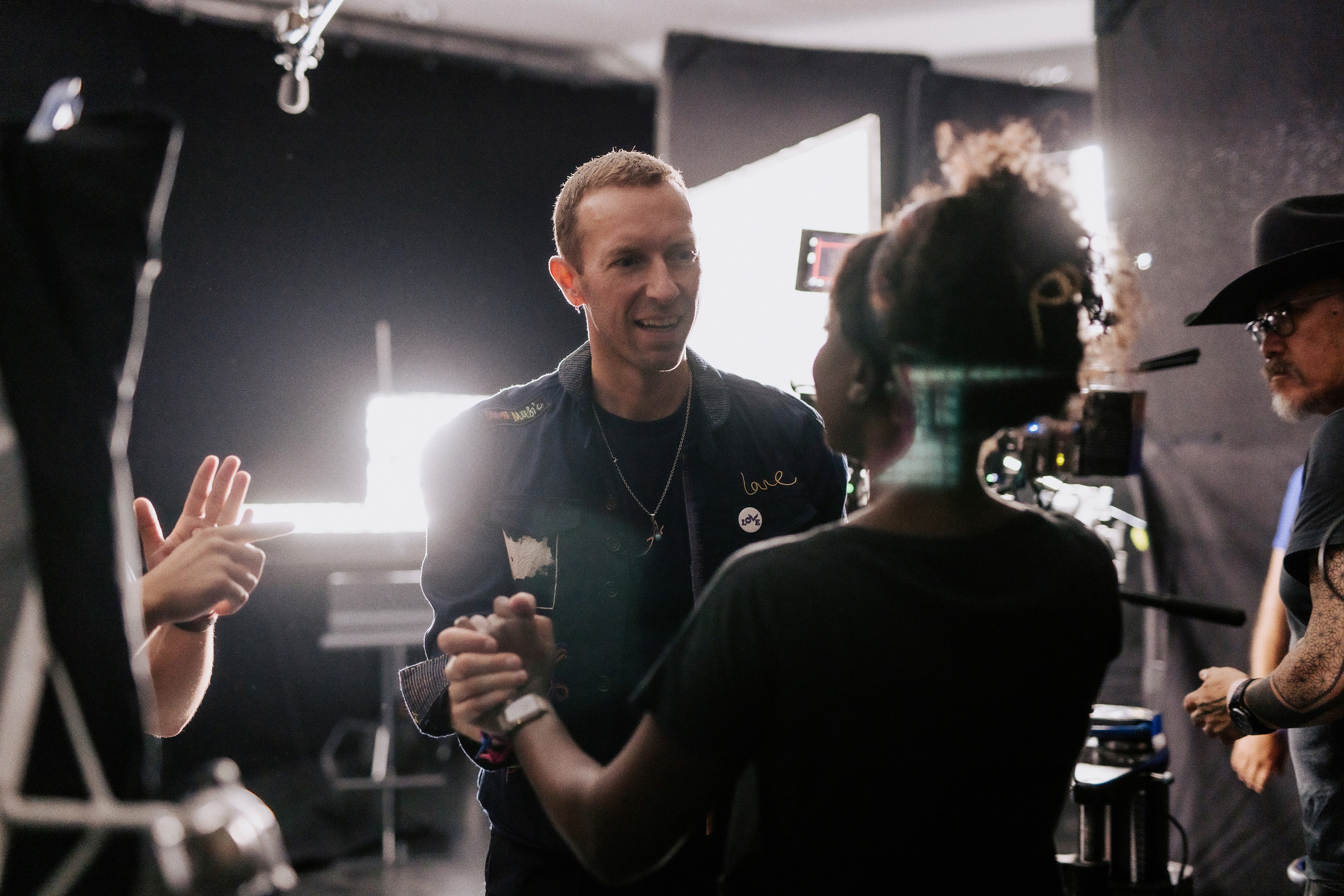 Chris Martin (a white, British man) and Natasha Ofili (a dark-skinned Black woman) are in a green room; Chris is holding Natasha’s hands while slightly smiling, and Natasha is looking at Chris as he talks. 