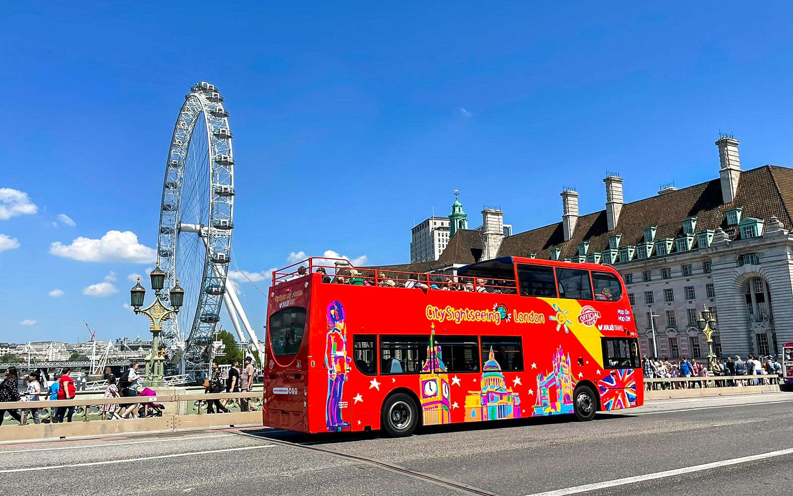 Red double-decker bus near the London Eye on a sunny day.