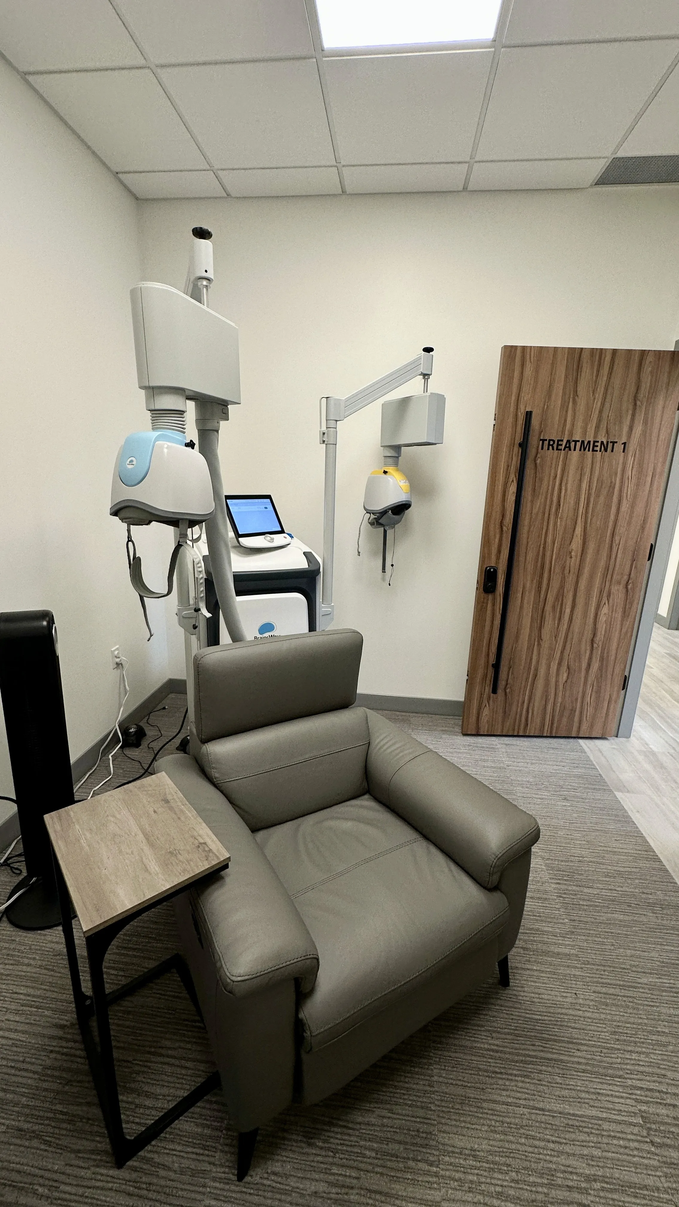 Deep TMS treatment device with coil beside a leather recliner chair next to a door labeled Treatment 1