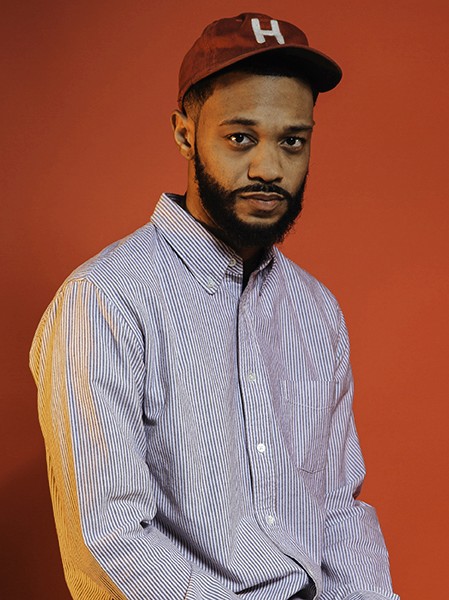 A man with dark skin and a beard wears a maroon baseball cap with a white "H" on the front. He wears a light blue and white vertically striped button-down shirt. The background is solid orange.