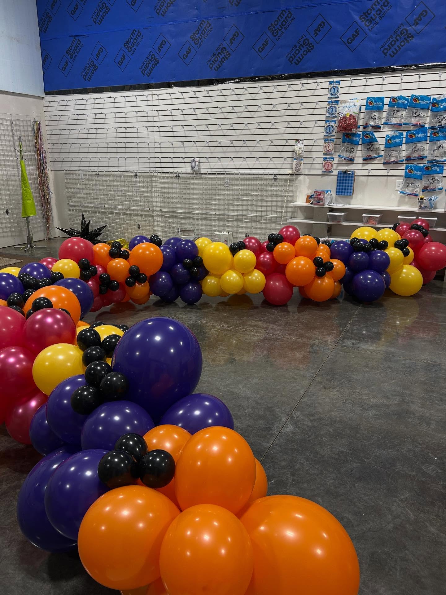 Colorful balloon garland with red, yellow, purple, and blue balloons arranged along a table at an indoor event by Be Bold Balloons