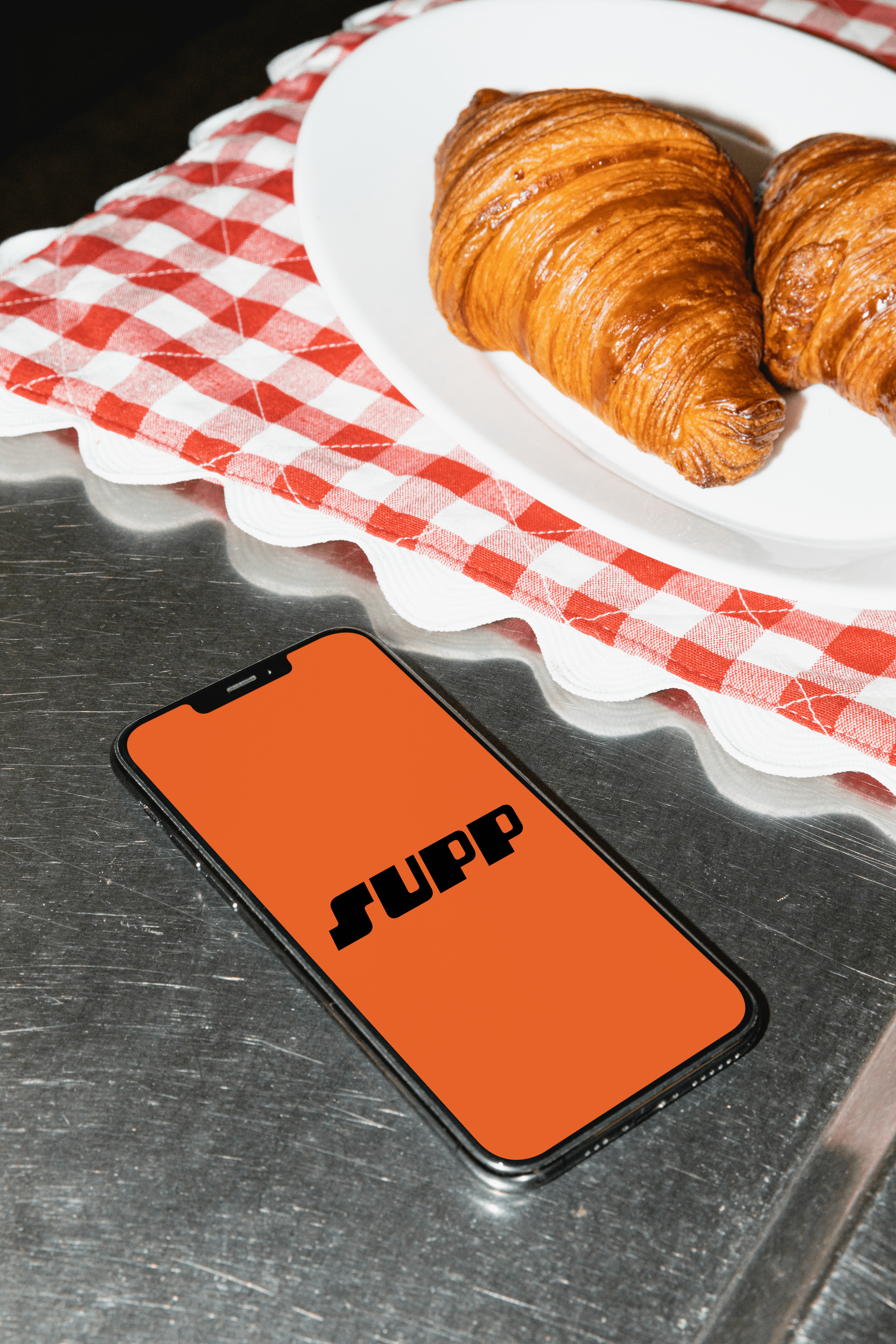 A phone displaying the Supp app logo on a cafe counter next to a plate of croissants