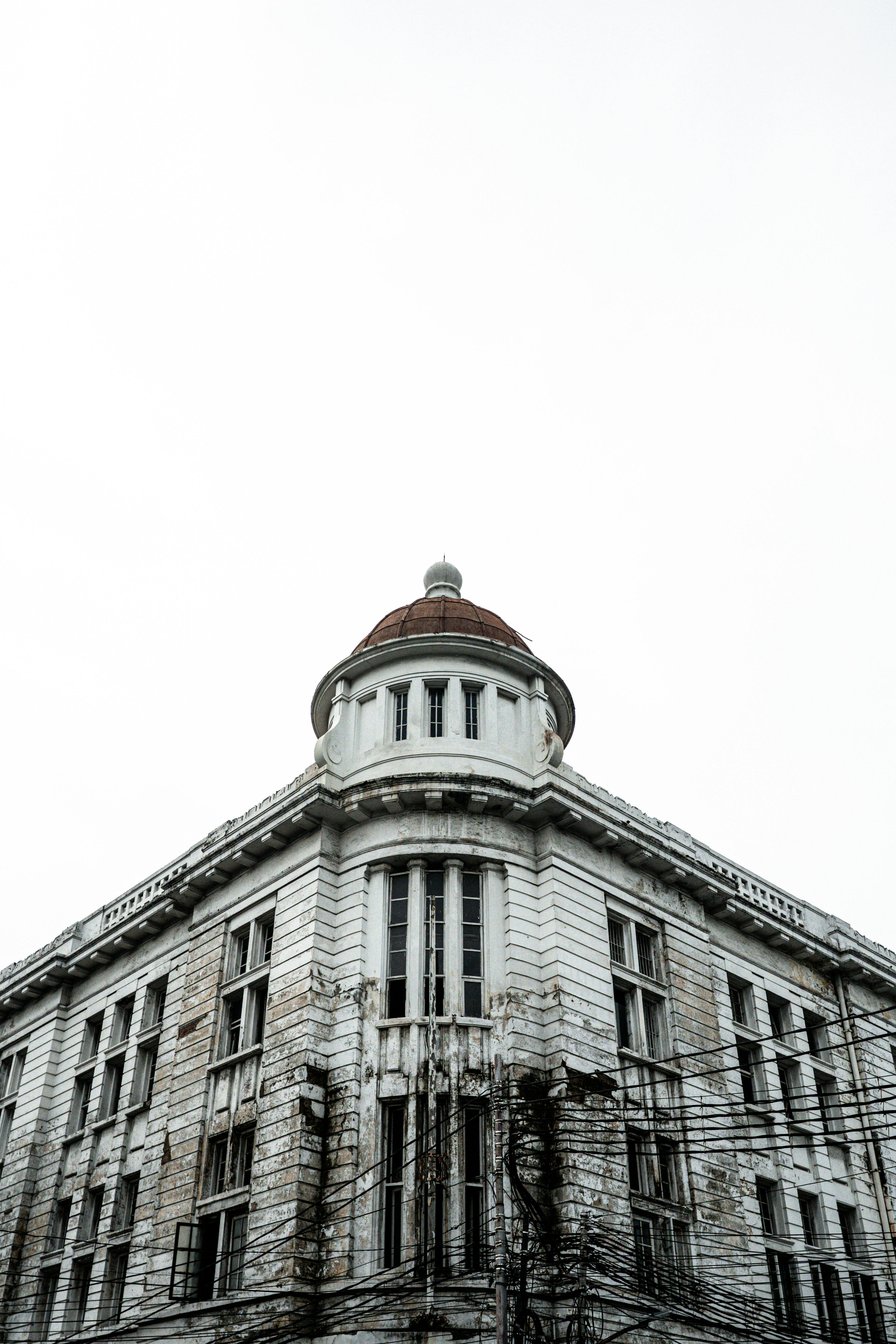 A historic, white colonial-style building with multiple windows and a prominent round tower featuring a red dome stands against a bright, overcast sky.