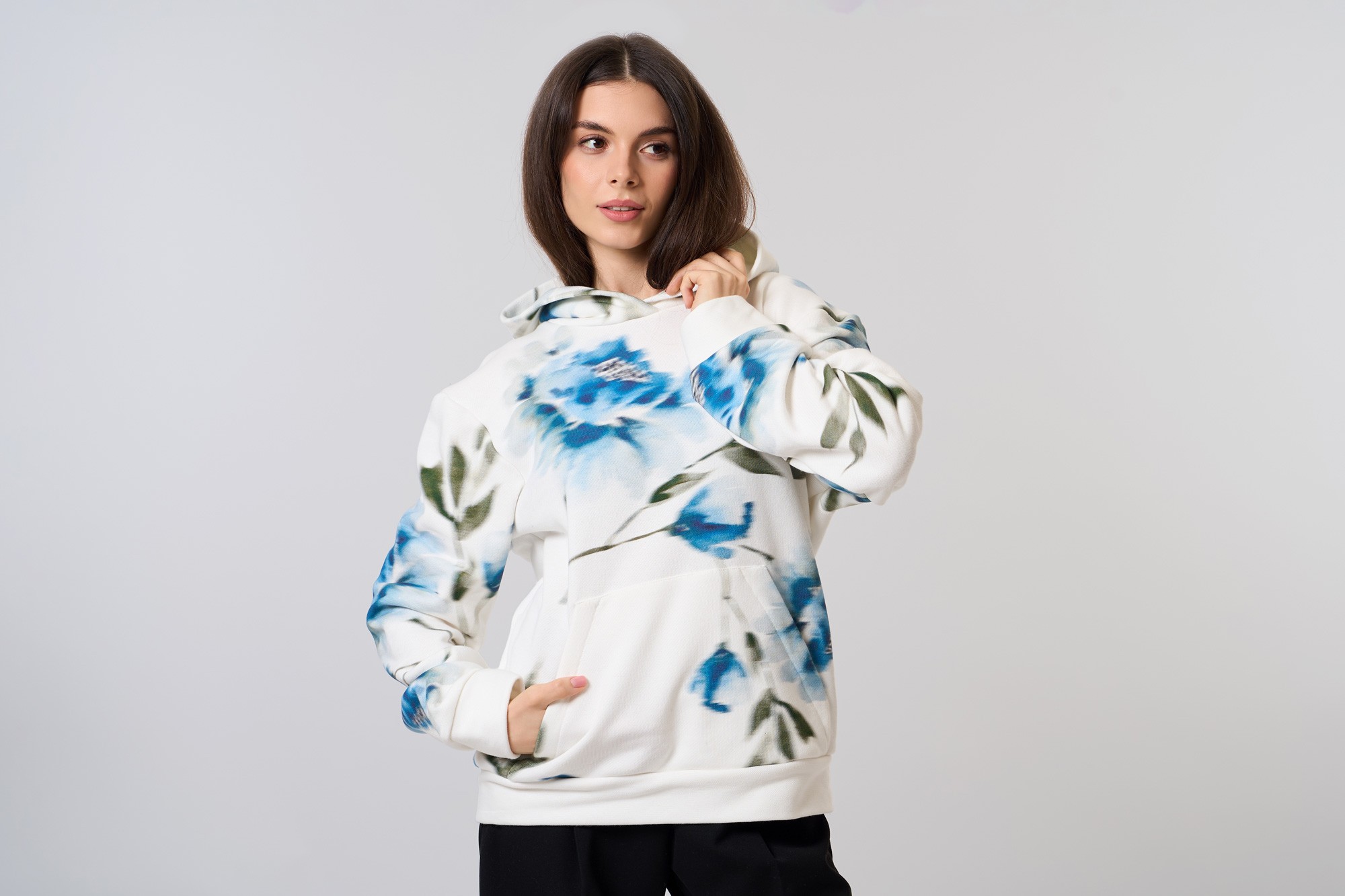 Sieviete baltā studijā valkā MUZE baltu hoodie ar zilu ziedu rakstu un roku ieliktu kabatā