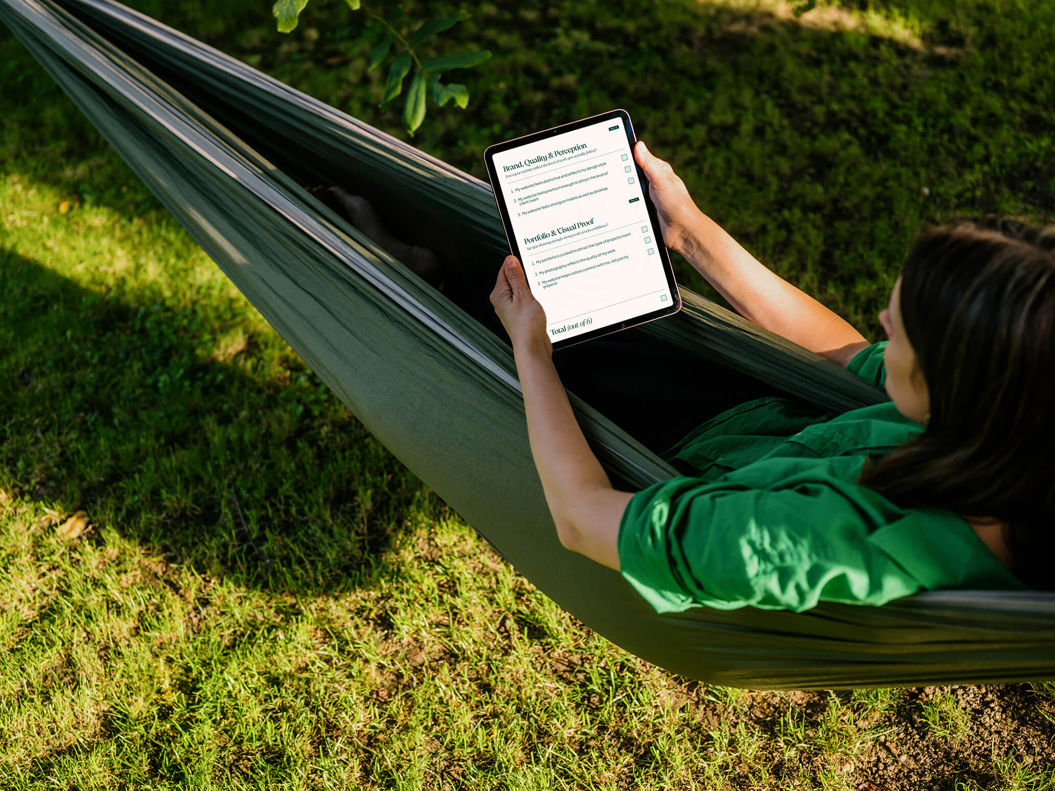 A person relaxes in a hammock, reading a an iPad outdoors on green grass in bright sunlight.