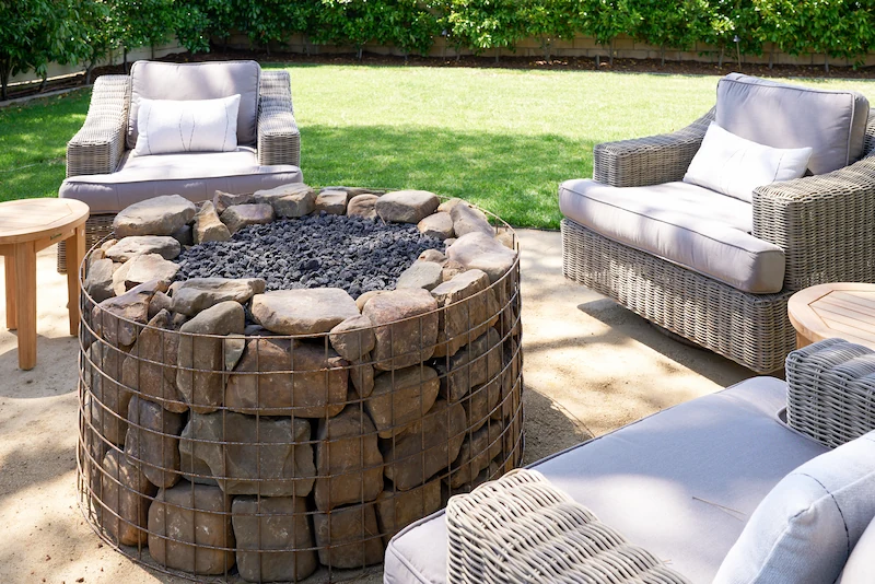 Inviting outdoor fire pit with stacked stone design surrounded by cozy wicker armchairs on lush grass. Costa Mesa Exterior Remodel by Todd Huge.