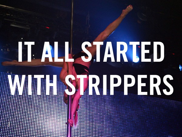 A woman in a black bra and shorts performs a pole dance with her arms extended upward and legs spread apart. She is positioned on a pole against a blue LED screen backdrop. Bright blue stage lighting illuminates the scene. Text overlaid on the image reads “IT ALL START WITH STRIPPERS”.