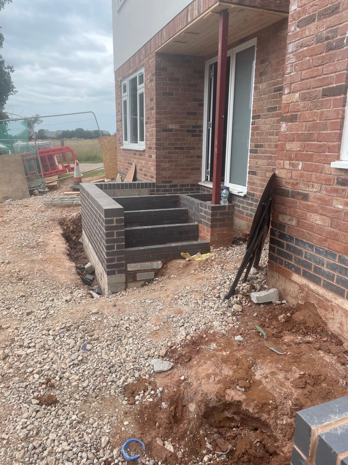 Construction area showing a building entrance with steps, gravel, and construction materials nearby under a cloudy sky.