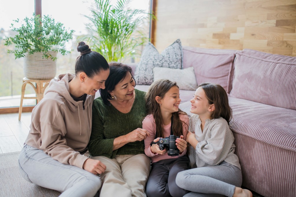 A multigenerational family shares a joyful moment in a cozy living room. The grandmother, mother, and two young girls sit together on the floor, smiling and looking at a camera one of the girls is holding. The warm and inviting setting, complete with comfortable seating and houseplants, highlights family bonding, storytelling, and the importance of capturing memories.