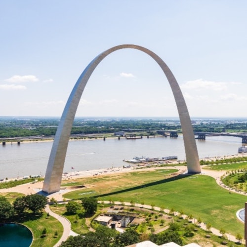 A large arch structure by a river, with parks, paths, and a bridge in the background under a clear, blue sky.