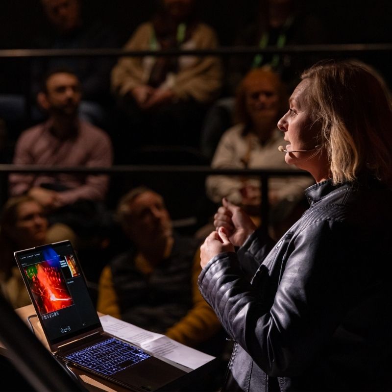 A womanA woman wearing a headset microphone speaks to an audience in a dimly lit showroom, presenting at an Innovation Week event at Lava Show in Reykjavík. wearing a headset microphone speaks to an audience in a dimly lit showroom, presenting at an Innovation Week event at Lava Show in Reykjavík.