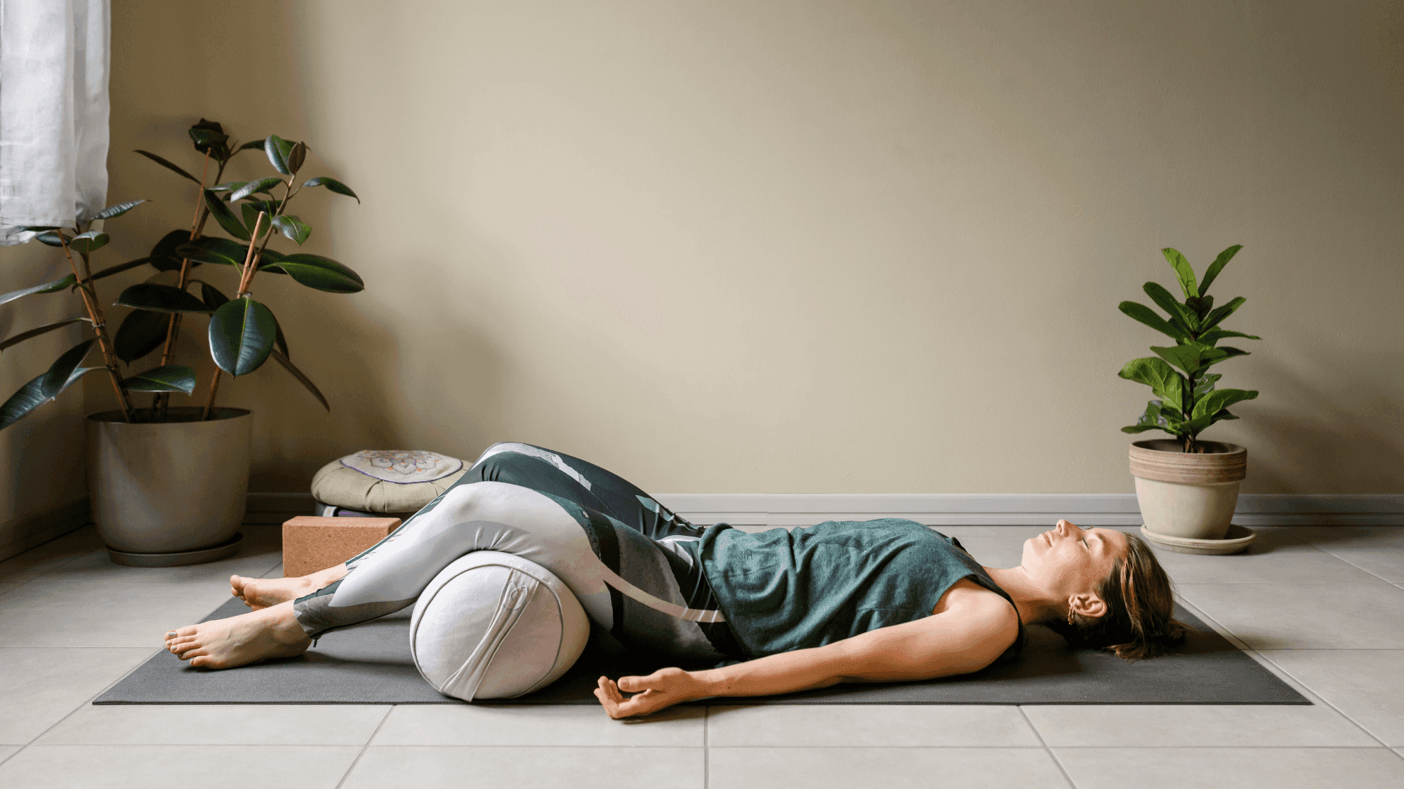 Woman practicing Supported Reclined Bound Angle Pose (Supta Baddha Konasana) on yoga mat with bolster, demonstrating restorative yoga in calm home setting with houseplants