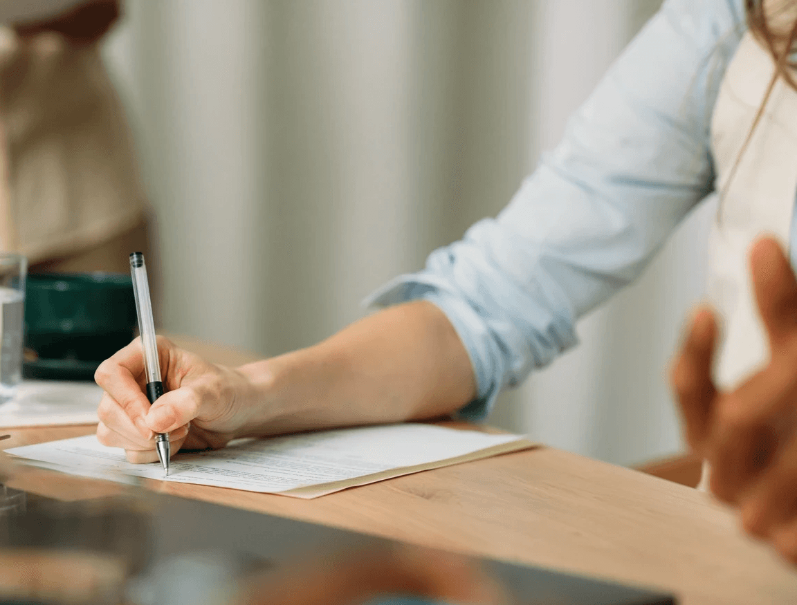 Close-up of a person taking notes during a business meeting, with colleagues, laptops and documents around a conference table.