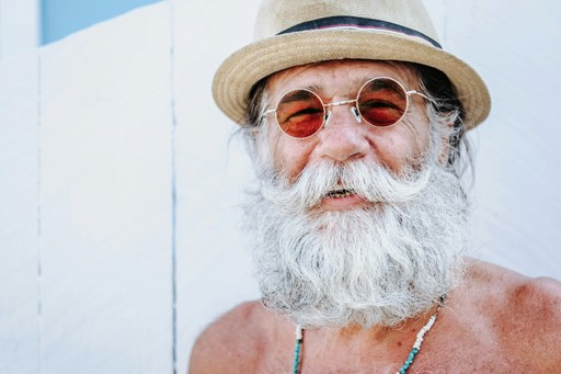 An older man with a long white beard and sunglasses, wearing a straw hat, stands against a light-colored background.